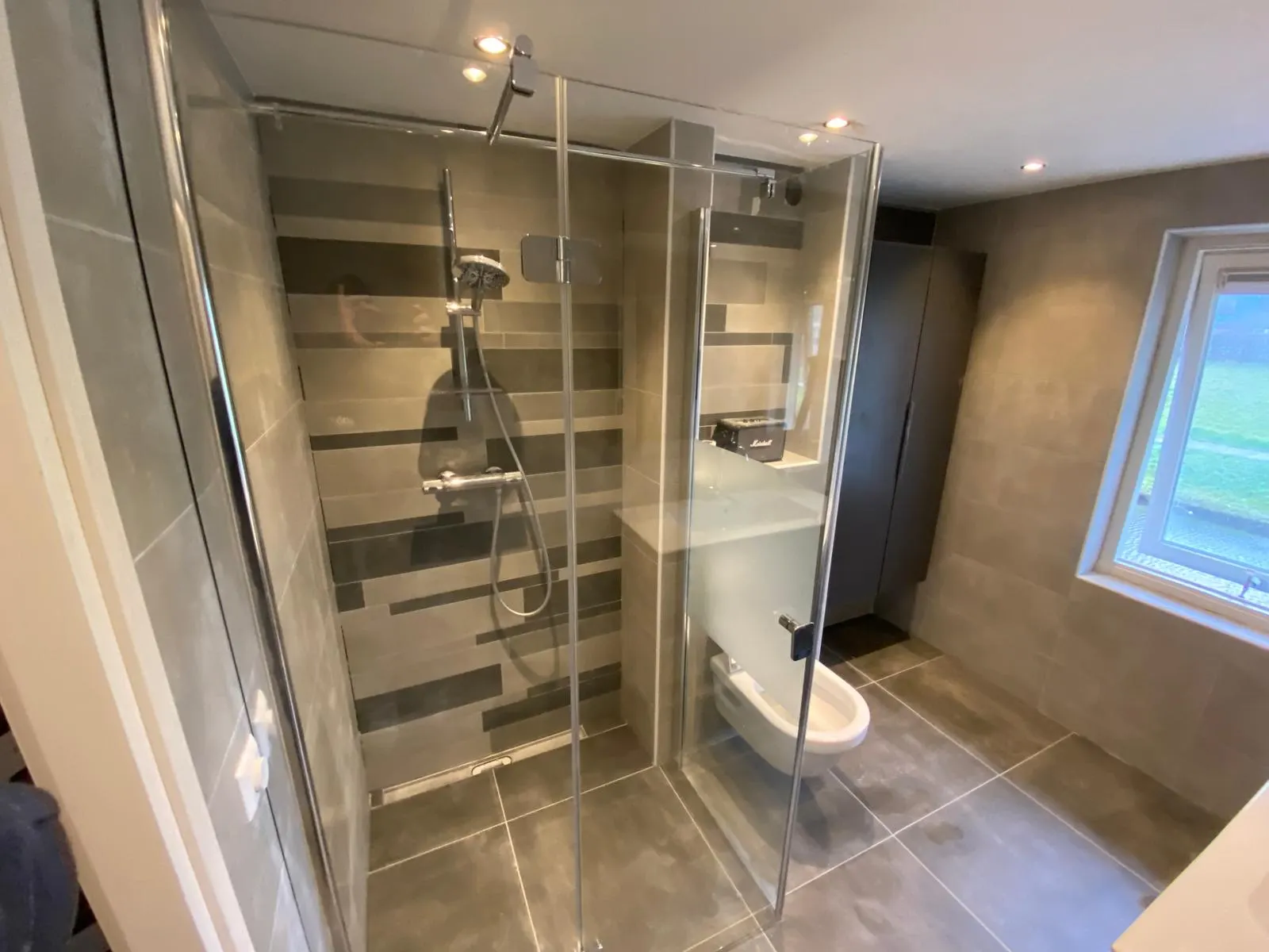 Gerenoveerde badkamer met grote grijze tegels, inloopdouche met streepaccent, douchegoot en hangend toilet