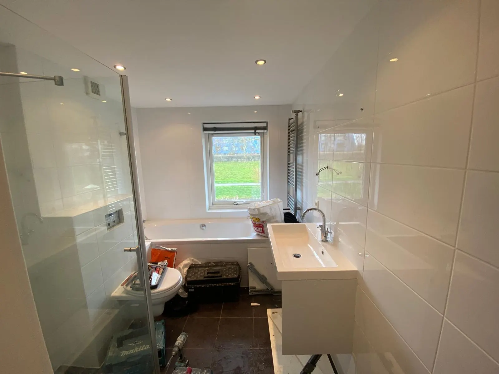 Voor: Witte betegelde badkamer met ligbad onder het raam, gereedschap op de vloer bij de start van de renovatie in Delft