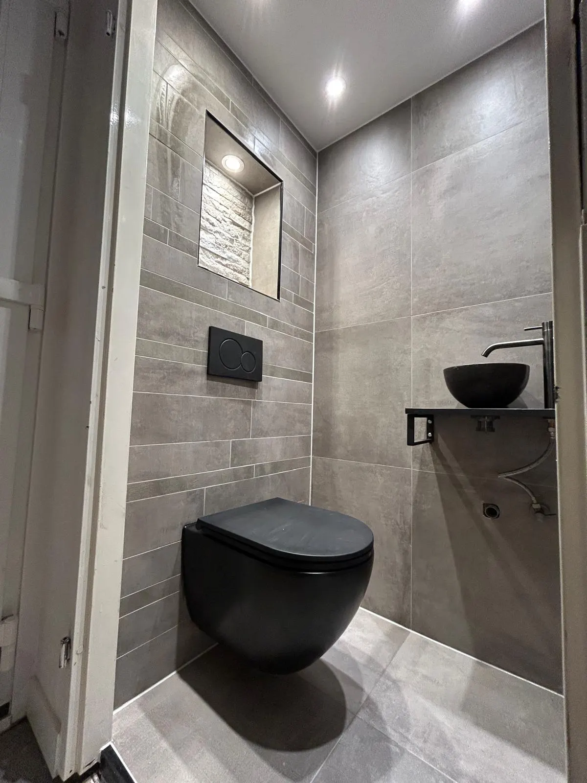 Gerenoveerd toilet in Delft met mat zwart hangend toilet, verlichte nis, grijze tegels en zwarte waskom op een plank